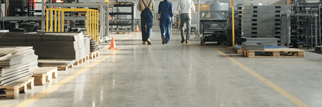 resin-flooring-1-v1 Three workers in safety gear walk away through an industrial facility with materials and machinery on either side.